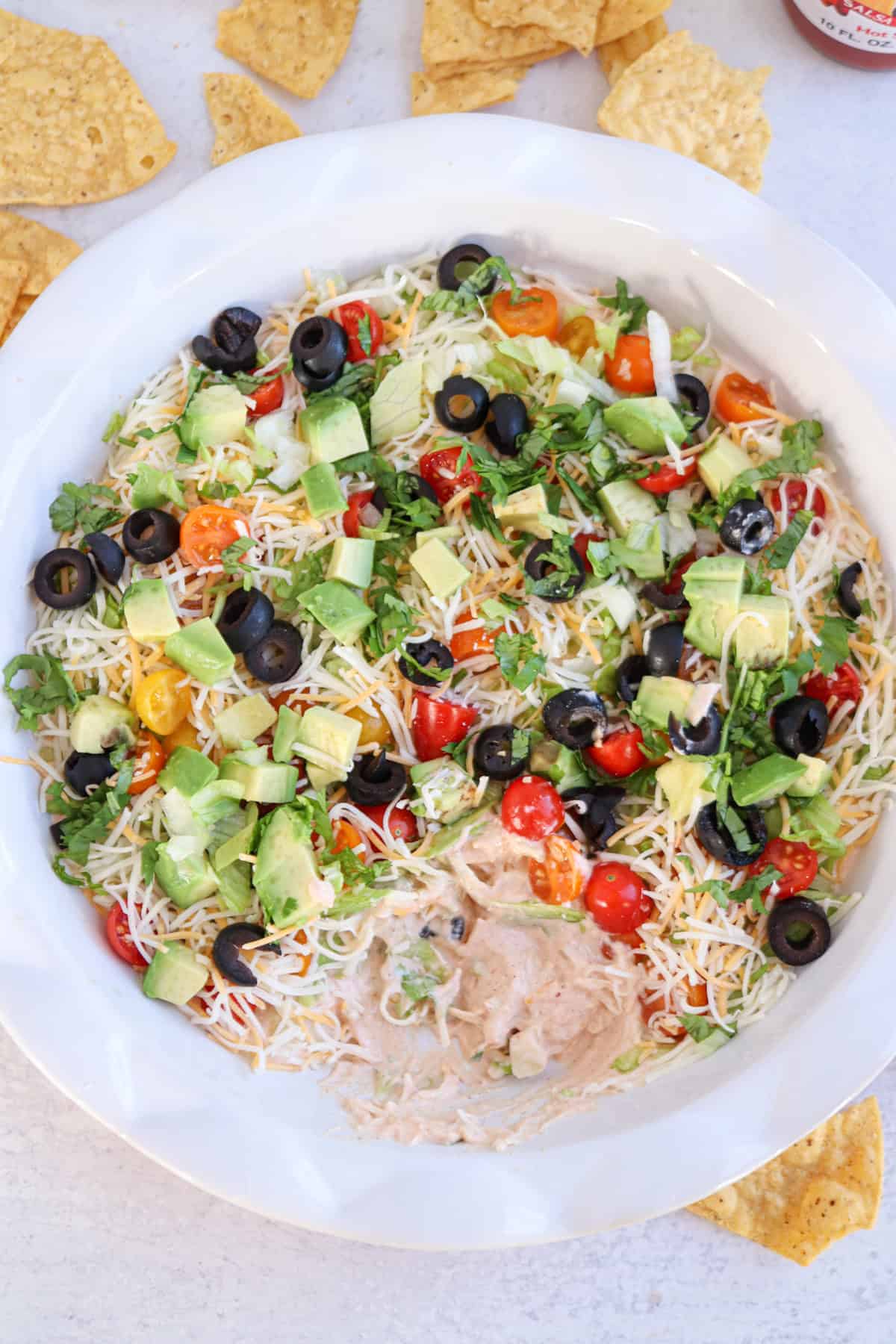 A colorful seven-layer dip in a white bowl, featuring layers of cheese, black olives, diced tomatoes, chopped avocado, and cilantro mixed with cottage cheese taco dip, surrounded by tortilla chips on a light surface.