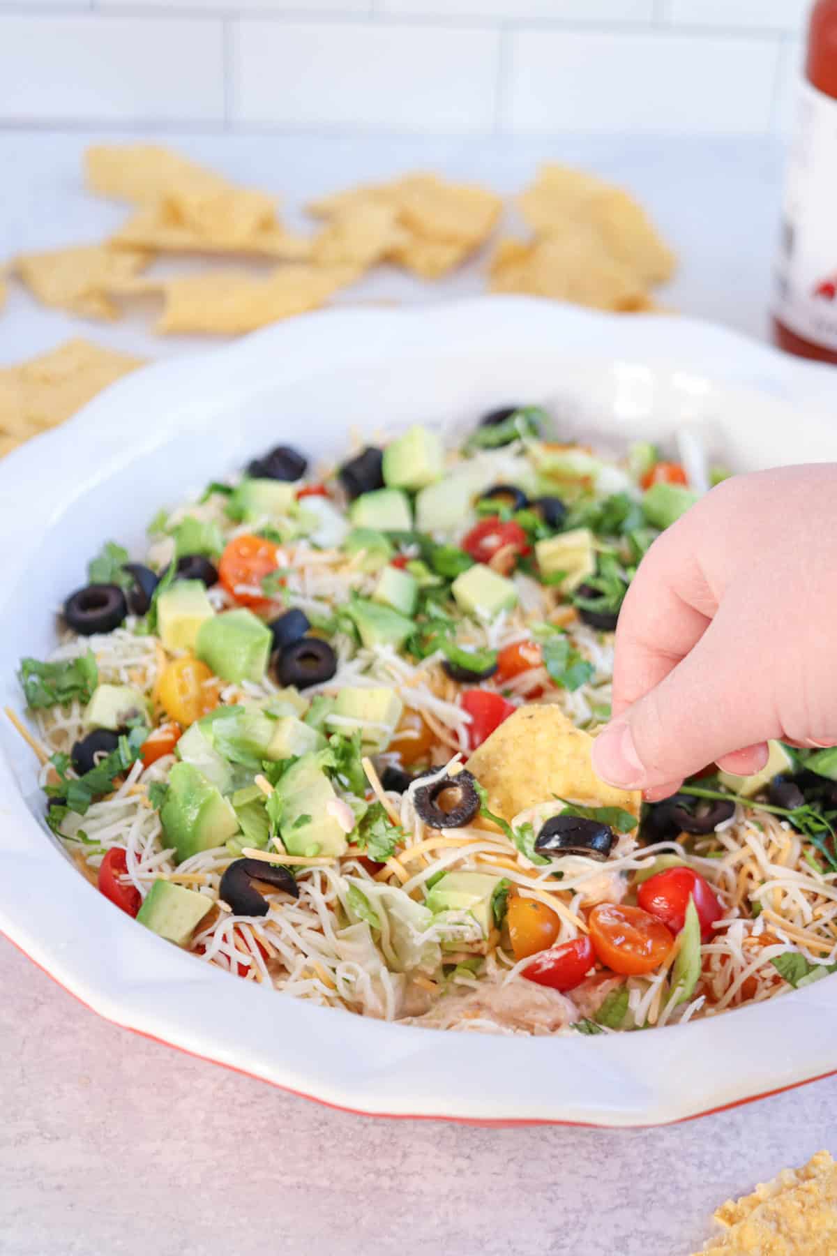 A hand dips a tortilla chip into a vibrant cottage cheese taco dip layered with chopped tomatoes, avocado, olives, shredded cheese, and cilantro in a white dish. Scattered tortilla chips linger in the background.