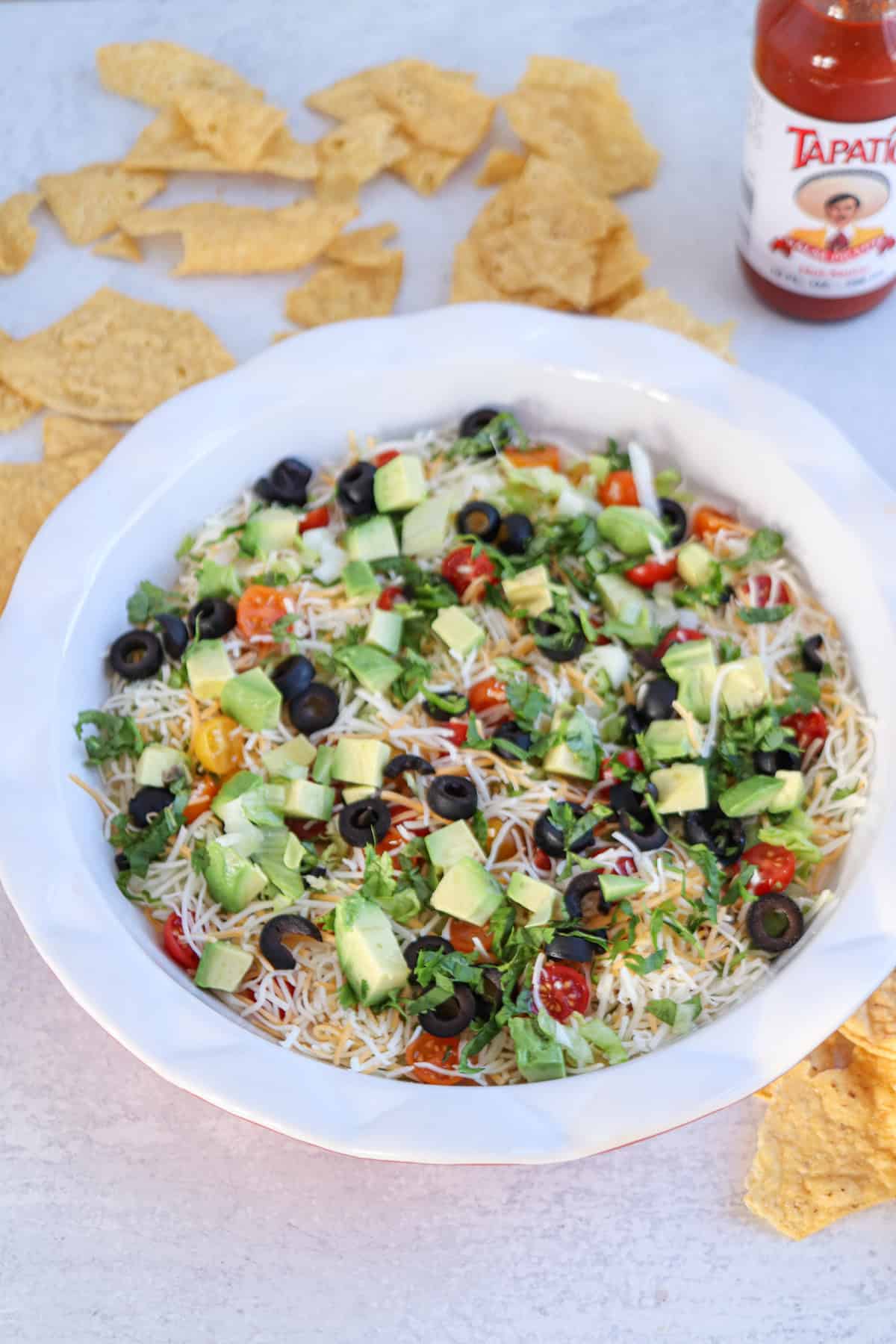 A round dish features a layered taco dip with cottage cheese, shredded cheese, black olives, avocado chunks, lettuce, and tomatoes. Tortilla chips and a bottle of hot sauce sit nearby on a light surface.