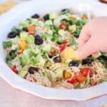 A hand dips a tortilla chip into a vibrant cottage cheese taco dip. The dish features avocado cubes, black olives, cherry tomatoes, shredded cheese, and fresh greens, all beautifully layered in a white serving dish.