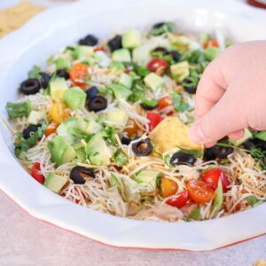 A hand dips a tortilla chip into a vibrant cottage cheese taco dip. The dish features avocado cubes, black olives, cherry tomatoes, shredded cheese, and fresh greens, all beautifully layered in a white serving dish.