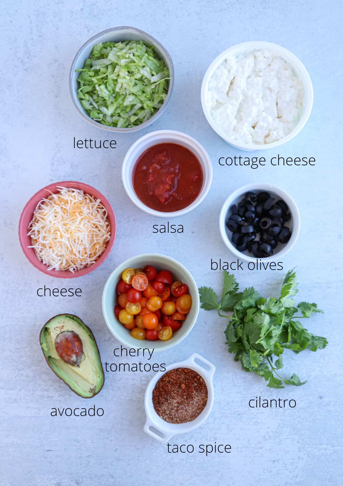 Top-down view of taco dip ingredients, featuring lettuce, salsa, cottage cheese, black olives, shredded cheese, cherry tomatoes, avocado, taco spice, and cilantro. Each component is neatly labeled in separate bowls on a light surface.