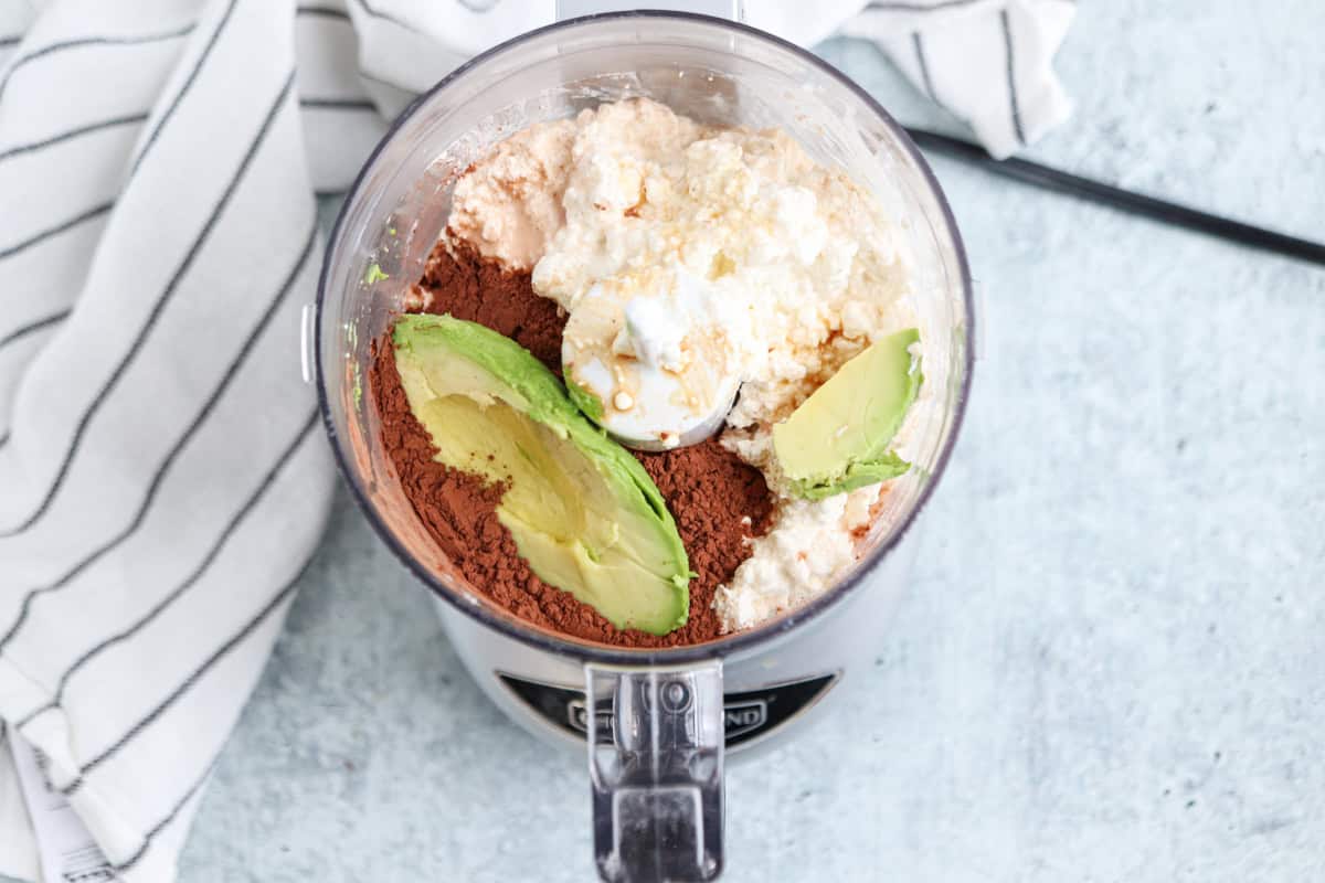 A food processor bowl contains avocado halves, cocoa powder, protein powder, and a creamy substance. A black and white striped cloth is partially visible on the side. The setting is a light-colored countertop.