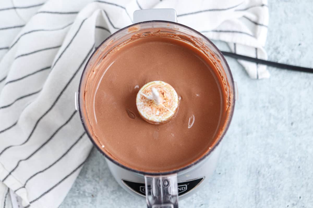 A food processor filled with smooth, creamy chocolate mixture sits on a light blue countertop. A white, striped kitchen towel is partially visible in the background.