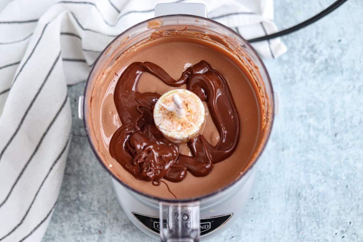 A food processor filled with smooth, creamy chocolate mixture. The surface has an uneven drizzle of melted chocolate. A striped cloth is partially visible beside the processor. The background is a light textured surface.