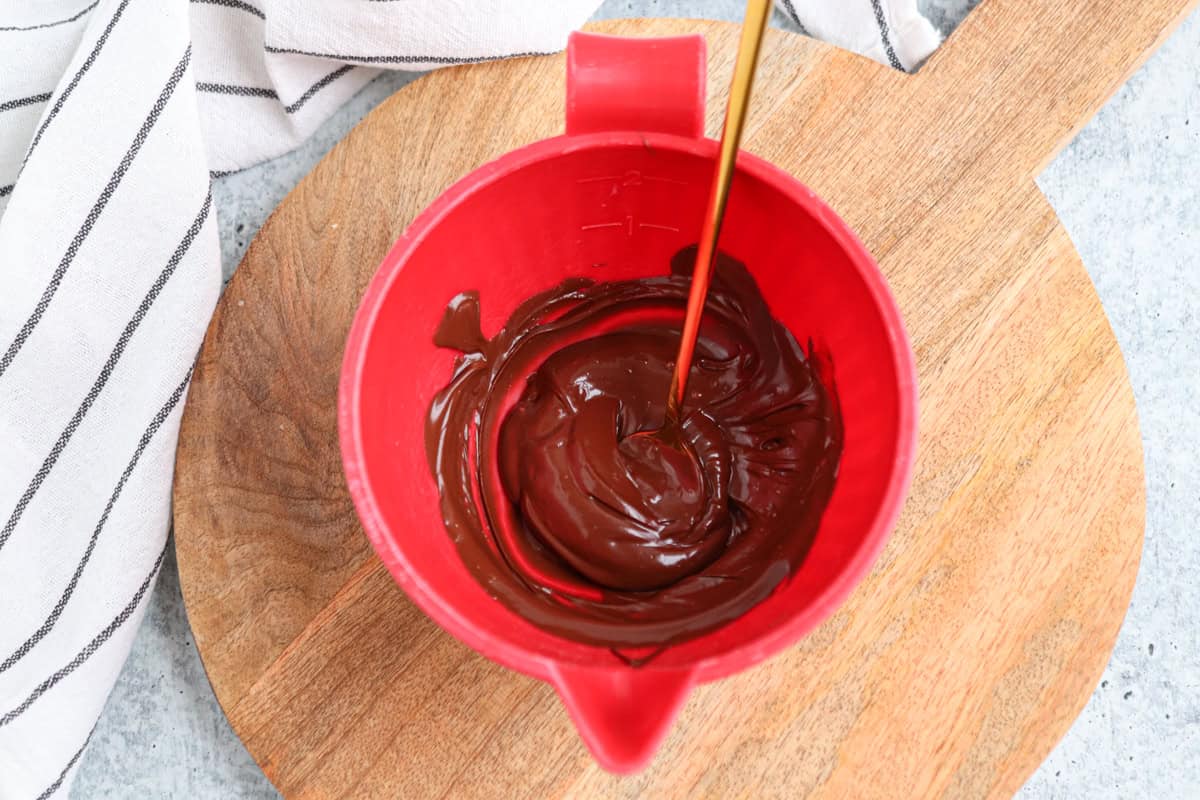 A red bowl with melted chocolate being stirred by a metal spoon, placed on a round wooden board. A white cloth with black stripes is seen on the side against a light gray surface.