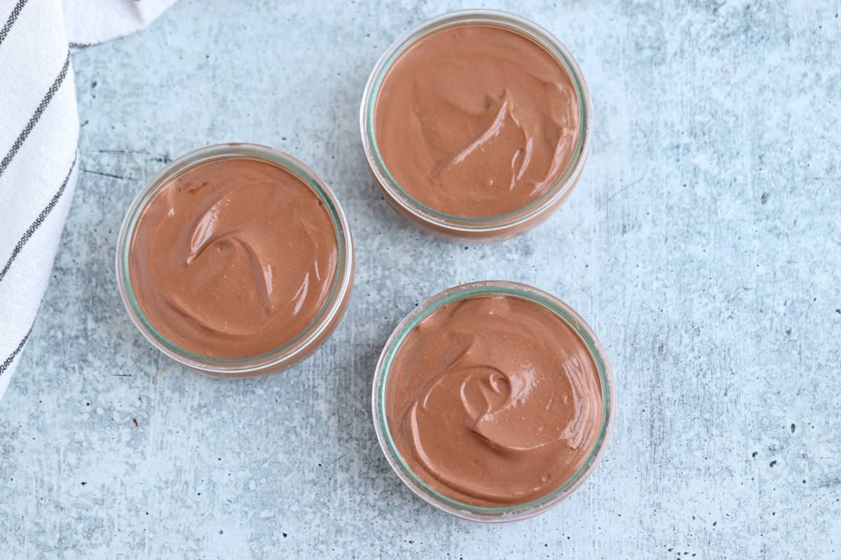 Three glass jars filled with creamy chocolate mousse are arranged on a textured gray surface. A white cloth with black stripes is partially visible on the left side.
