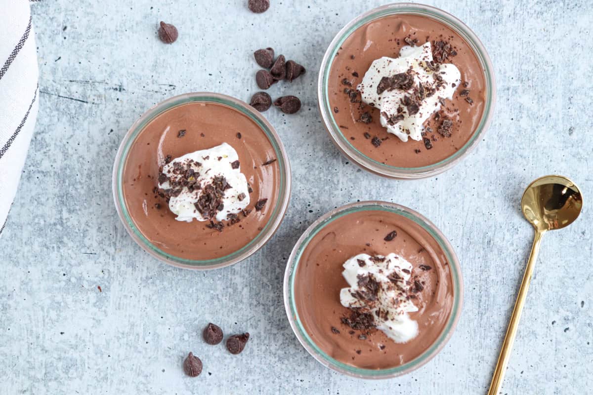 Three glass bowls of chocolate mousse topped with whipped cream and chocolate shavings are arranged on a light surface. Scattered chocolate chips and a gold spoon lie nearby. A corner of a striped cloth is visible.