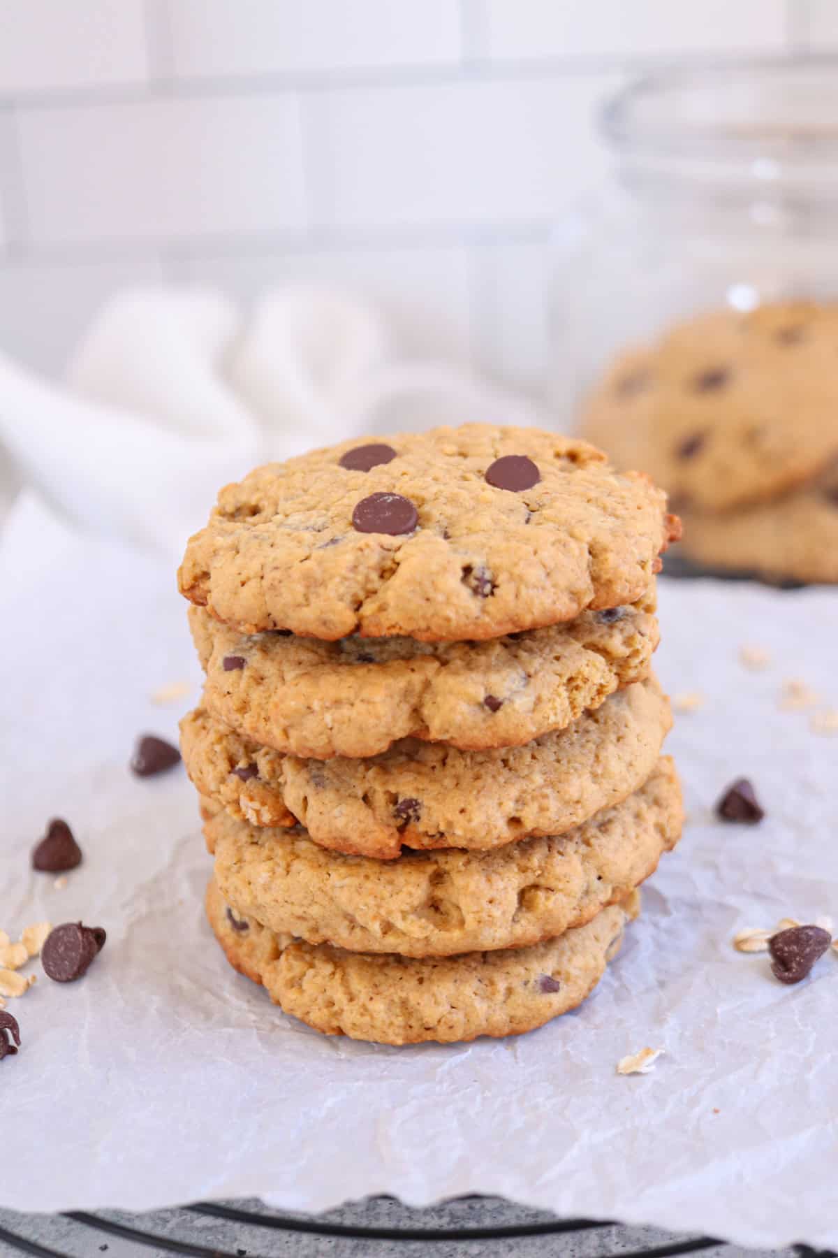 A stack of five chocolate chip cookies sits on a piece of white parchment paper. Some chocolate chips and crumbs are scattered around, adding to the rustic charm. A glass jar filled with more cookies is partially visible in the background.