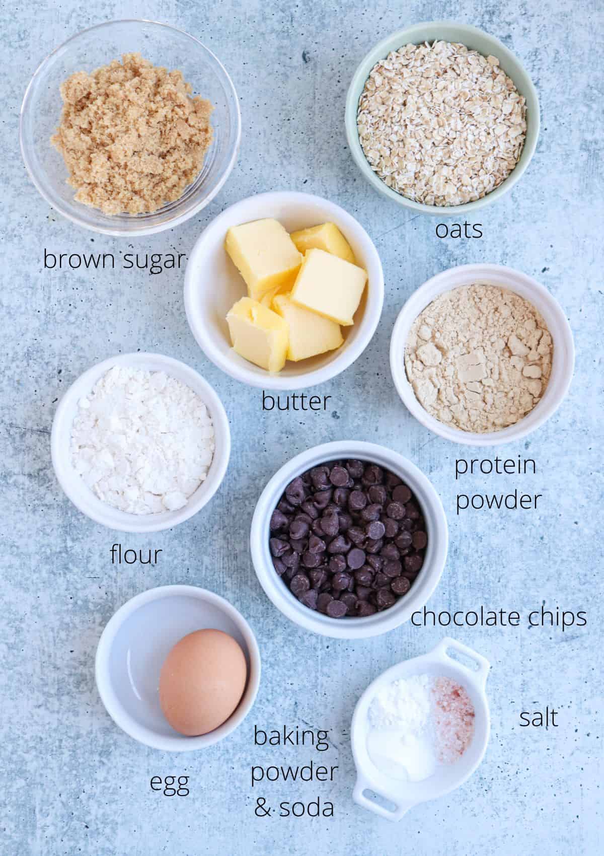 Top-down view of baking ingredients arranged in bowls on a countertop: brown sugar, oats, butter, protein powder, flour, chocolate chips, and an egg. Baking powder, baking soda, and salt are labeled alongside each item.