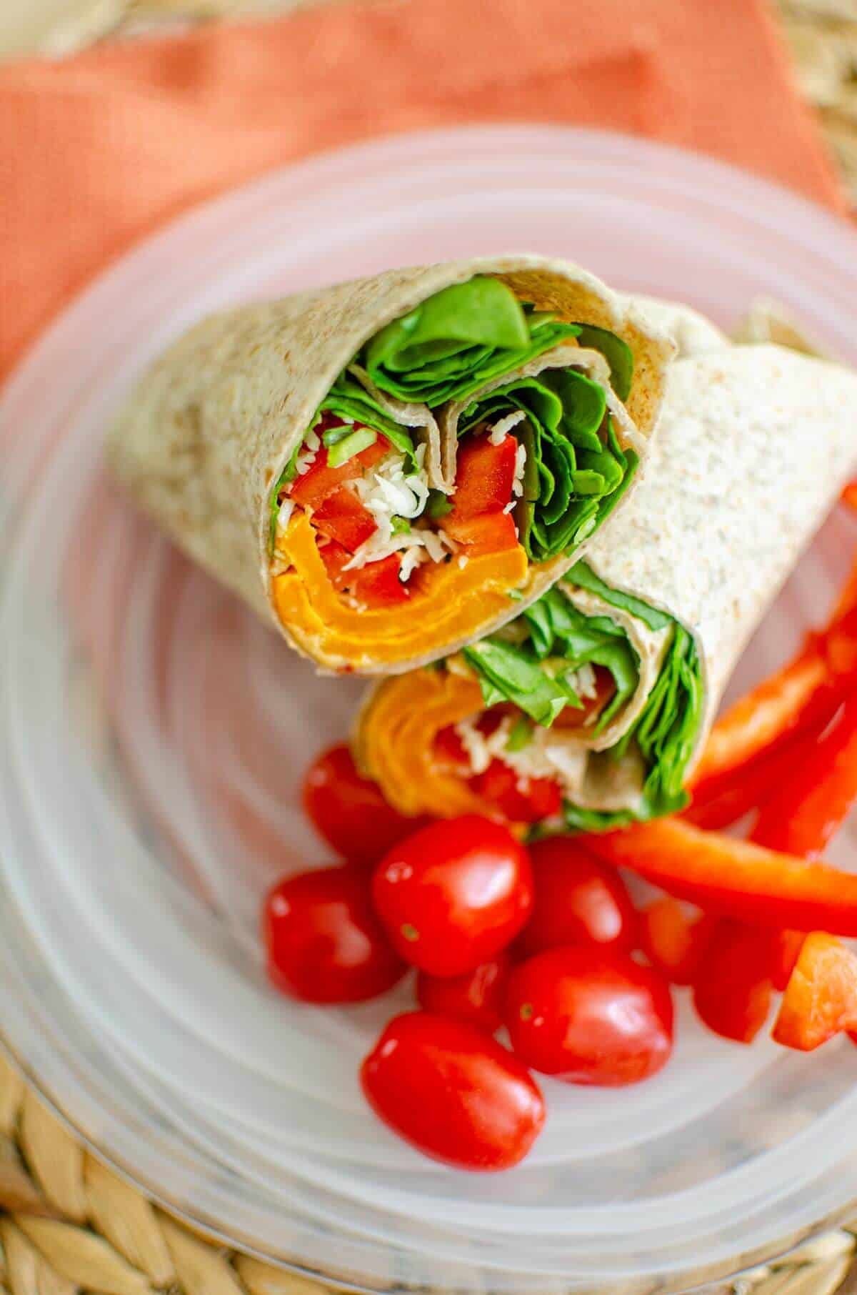 Two tortilla wraps filled with spinach, red bell peppers, carrots, and shredded cheese are placed on a clear plate. Cherry tomatoes and red bell pepper slices are arranged beside the wraps. A woven mat and an orange napkin are visible underneath.