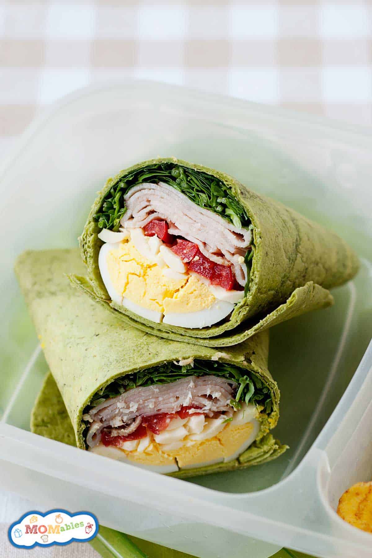 A spinach wrap cut in half, filled with boiled egg slices, turkey, spinach leaves, and red bell pepper strips, placed in a clear container. A colorful logo is visible in the corner of the image.