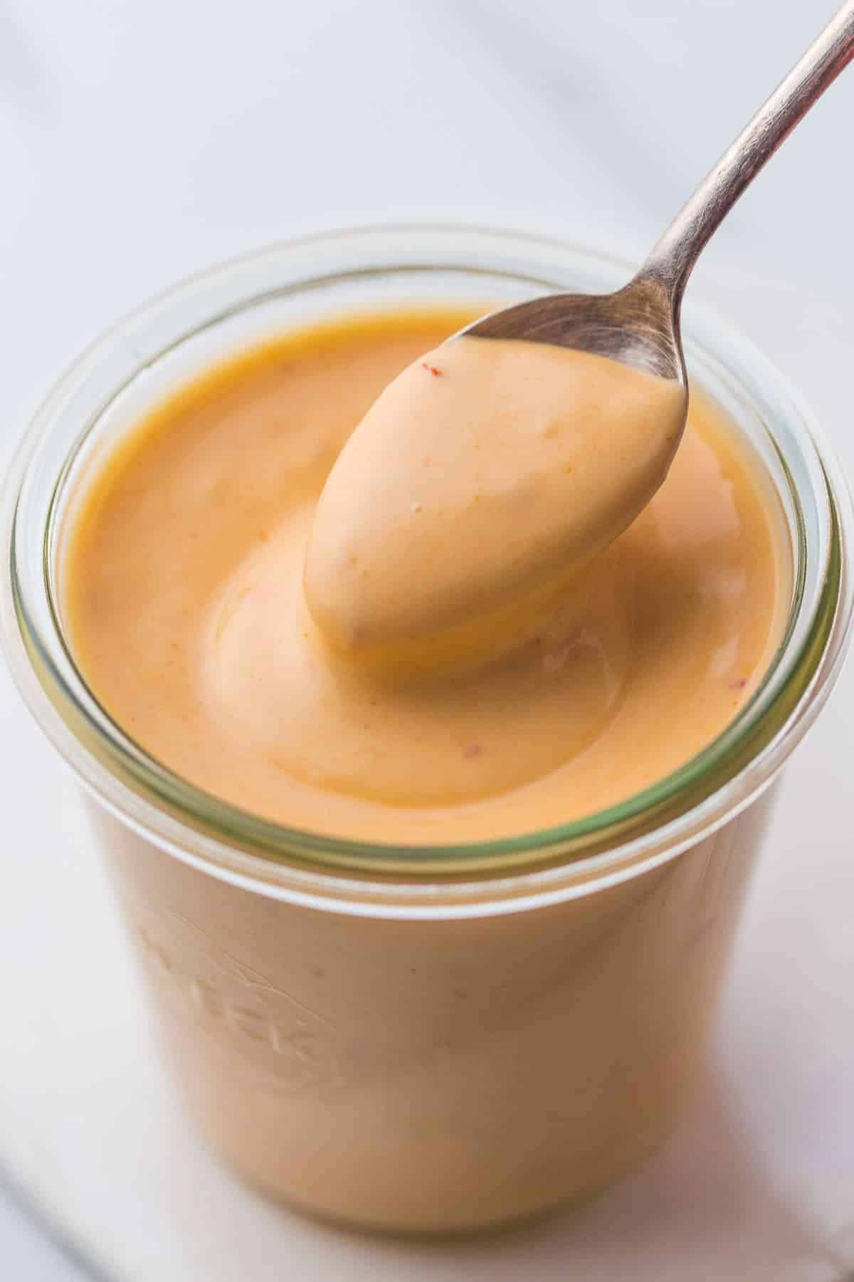 A spoon is dipped into a glass jar filled with creamy, light orange sauce. The sauce appears smooth and swirled, with hints of spices visible. The jar sits on a light-colored surface.