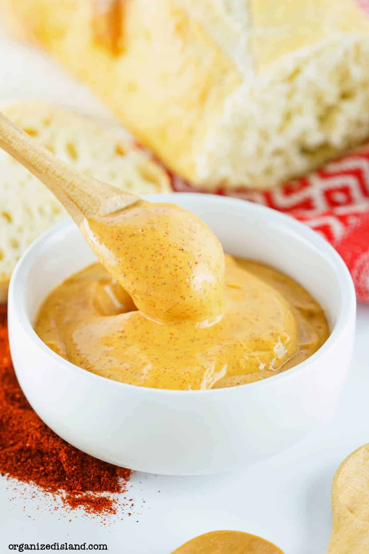 A wooden spoon dipping into a white bowl of creamy orange sauce, surrounded by a sprinkle of red spice. Part of a sliced bread loaf and a red and white cloth are visible in the background.