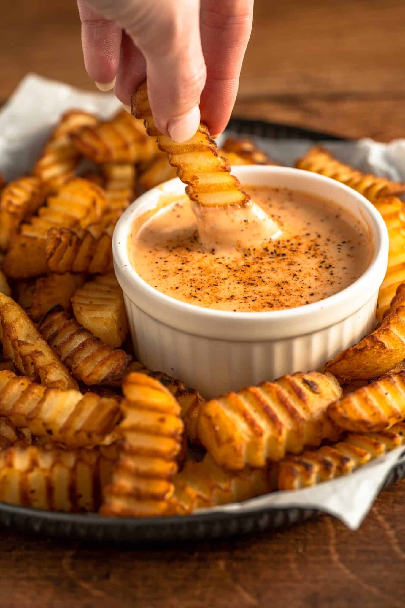 A hand dipping a crinkle-cut fry into a creamy, seasoned dipping sauce in a white ramekin, surrounded by more crinkle-cut fries on a dark plate. The fries are golden brown and crispy.