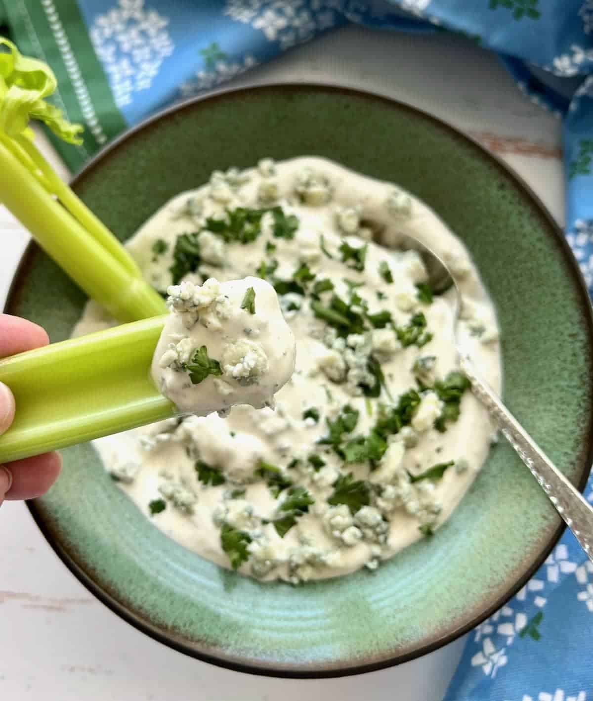 A green bowl filled with creamy blue cheese dressing, garnished with chopped herbs. A hand holds a celery stick dipped in the dressing. A silver spoon is placed in the bowl. A blue and white patterned cloth is in the background.