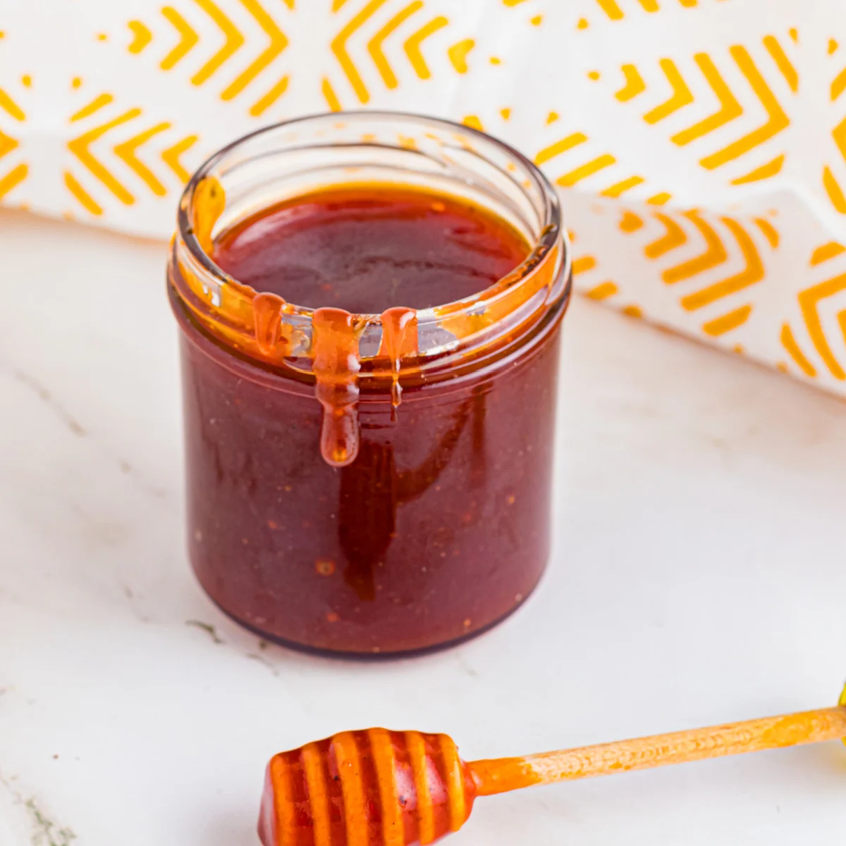 A jar of rich, dark sauce sits on a white surface. The sauce slightly drips down the side. A honey dipper coated with the same sauce lies nearby. The background features a white and yellow chevron-patterned cloth.