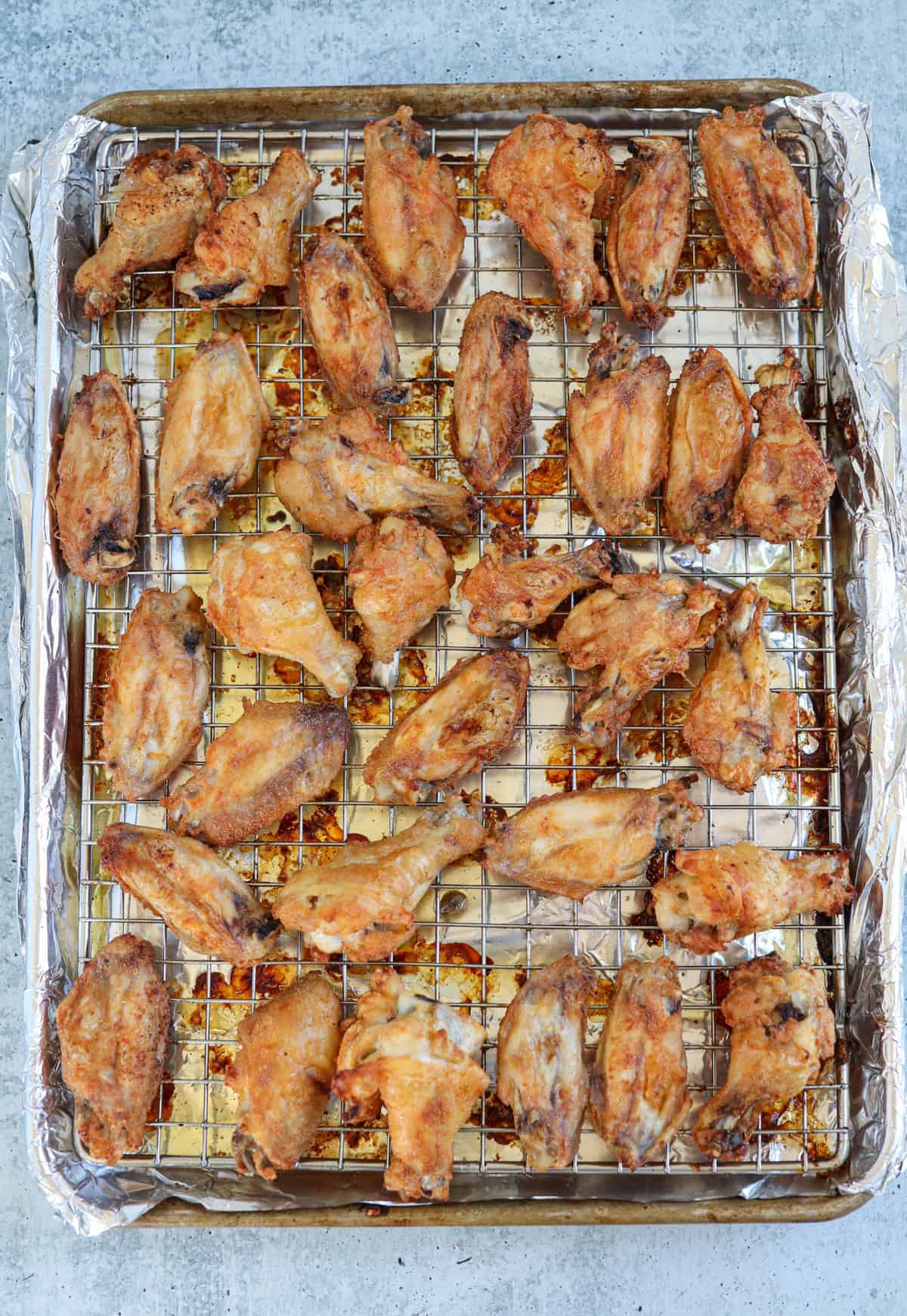 A tray lined with foil holds numerous golden-brown chicken wings arranged on a wire rack, freshly baked and evenly spaced. The wings appear crispy and well-cooked.