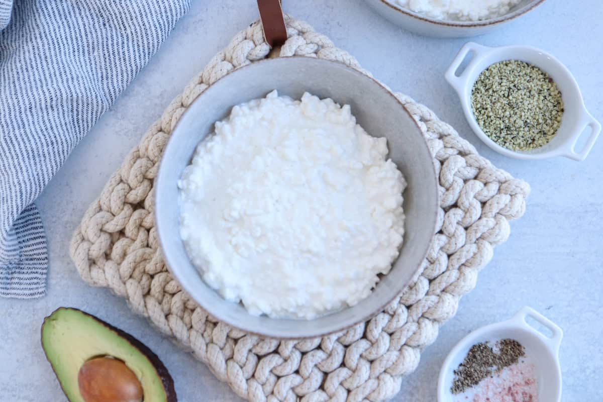 A bowl of cottage cheese sits on a knitted coaster. Next to it are half an avocado, a small dish of hemp seeds, and a dish containing salt and pepper. A striped cloth is partially visible on the side.