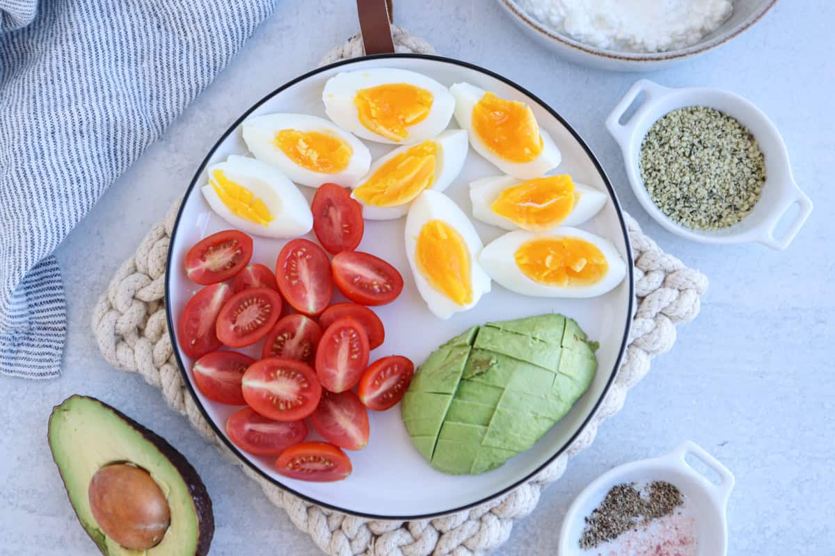 A plate with sliced boiled eggs, cherry tomatoes, and avocado. Surrounding the plate are small bowls of cottage cheese, hemp seeds, and salt with pepper. A halved avocado and a gray striped cloth are nearby on a textured gray surface.