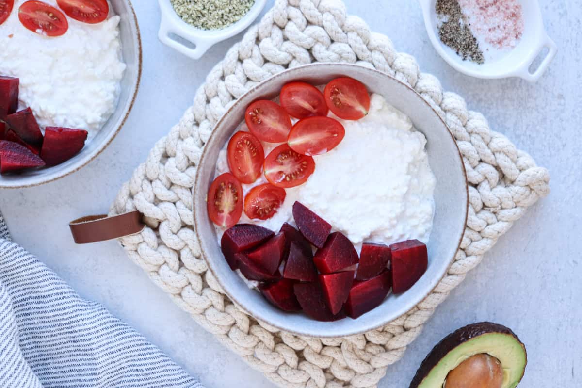 A bowl of cottage cheese topped with halved cherry tomatoes and sliced beets is placed on a knitted coaster. Nearby are small dishes of seasonings, a halved avocado, and a striped cloth. Another similar bowl is partially visible on the side.
