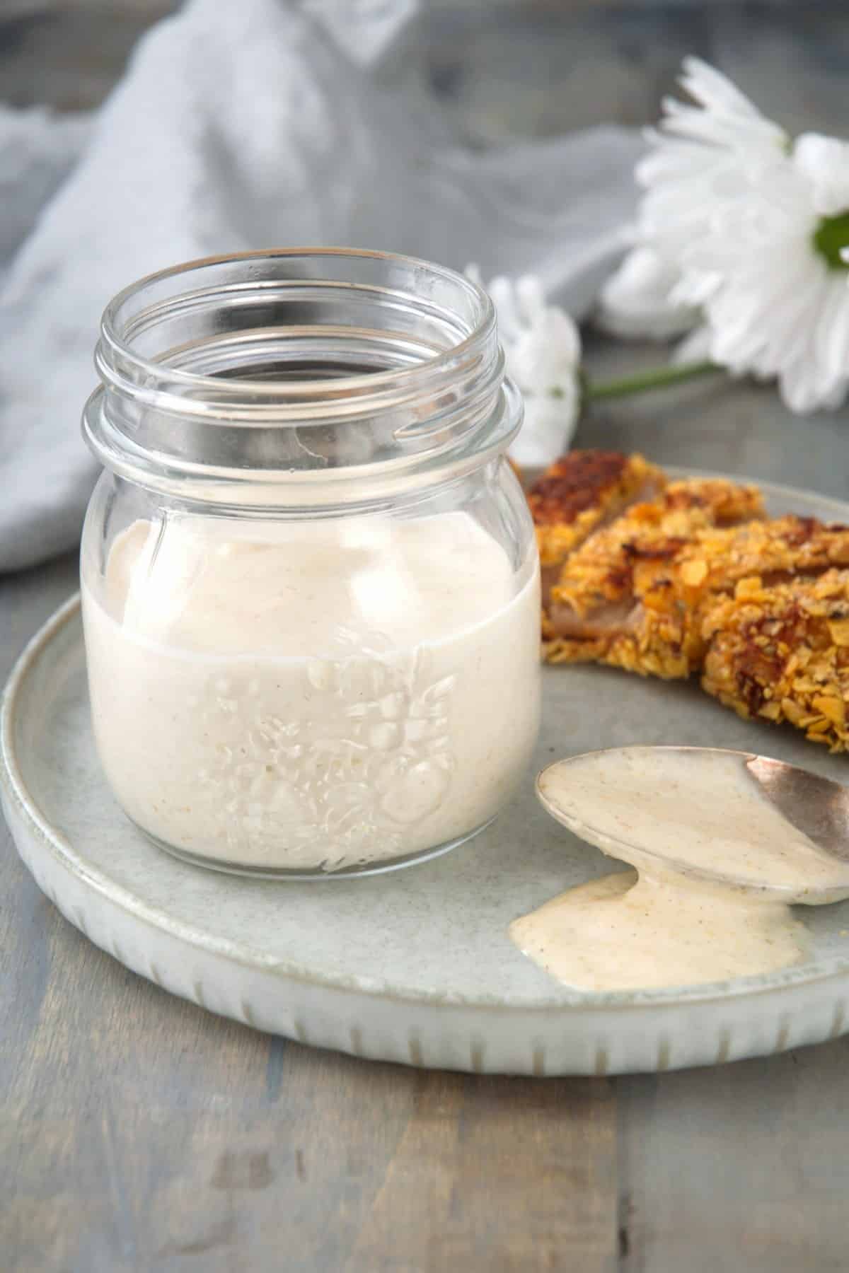 A small glass jar of creamy sauce sits on a gray plate with a spoon resting nearby, slightly dripped with the sauce. In the background, there is a piece of food crusted with a crispy coating and a white flower for decoration.
