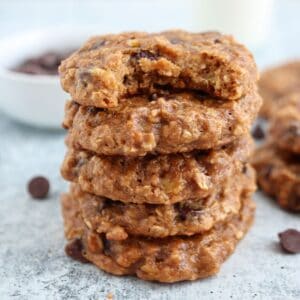 A stack of five chocolate chip oatmeal cookies sits on a textured surface. The top cookie has a bite taken out of it. A few chocolate chips are scattered around, and a bowl of more chips is blurred in the background.