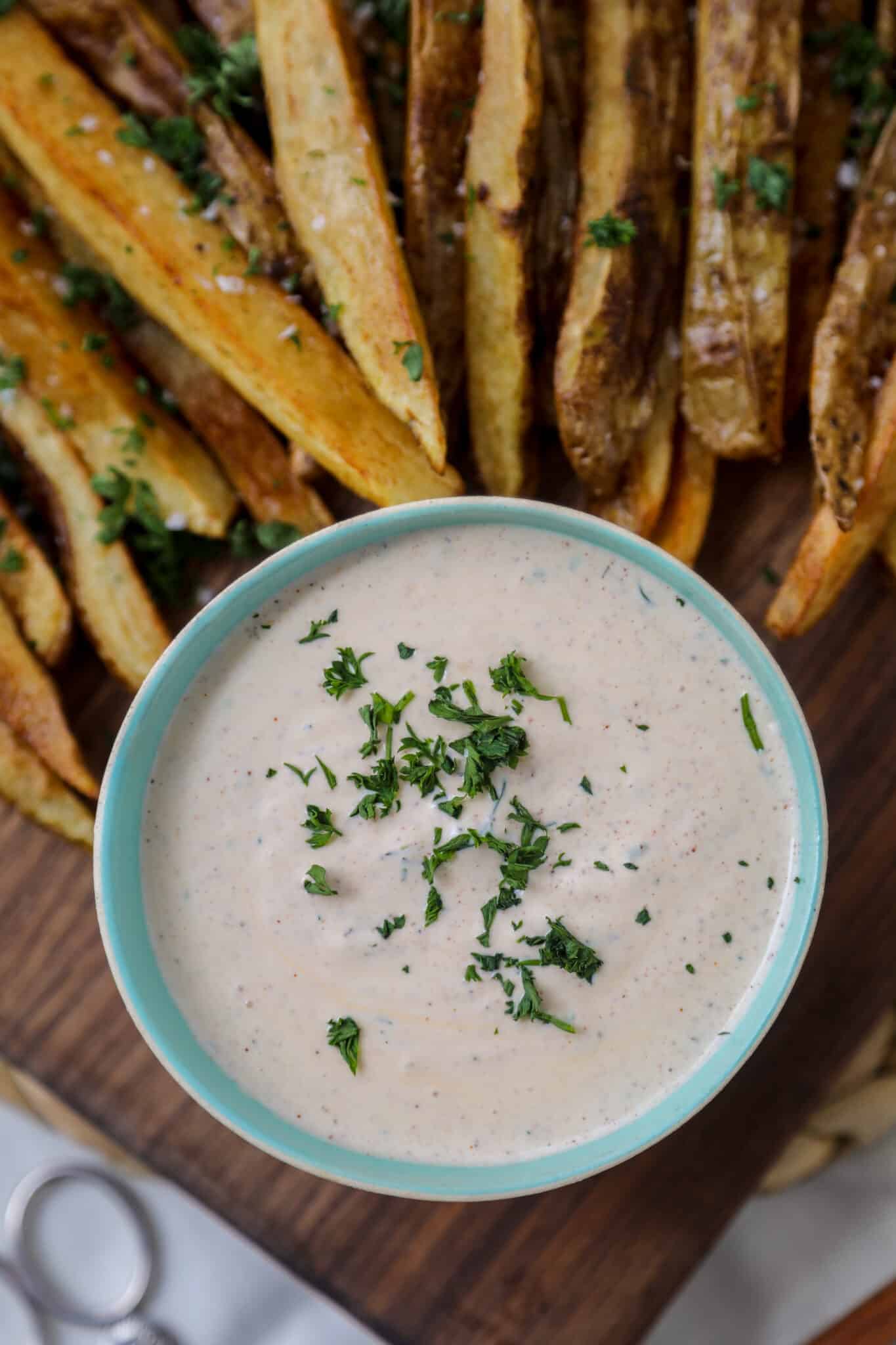 A bowl of creamy dipping sauce garnished with chopped herbs sits on a wooden board. Next to the bowl are golden-brown potato wedges sprinkled with herbs. The scene is inviting and freshly prepared.
