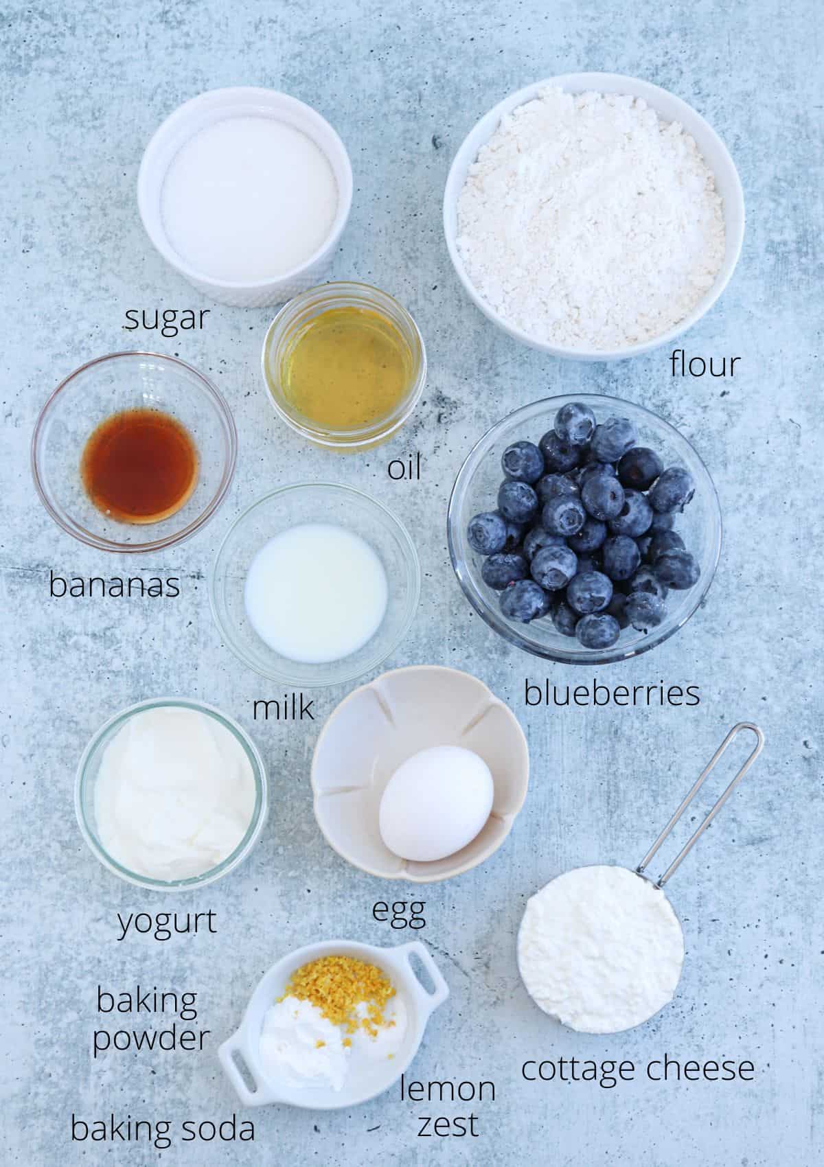 Various baking ingredients are arranged on a gray surface, each labeled: sugar, flour, bananas, oil, blueberries, vanilla, milk, yogurt, egg, baking powder, baking soda, lemon zest, and cottage cheese.