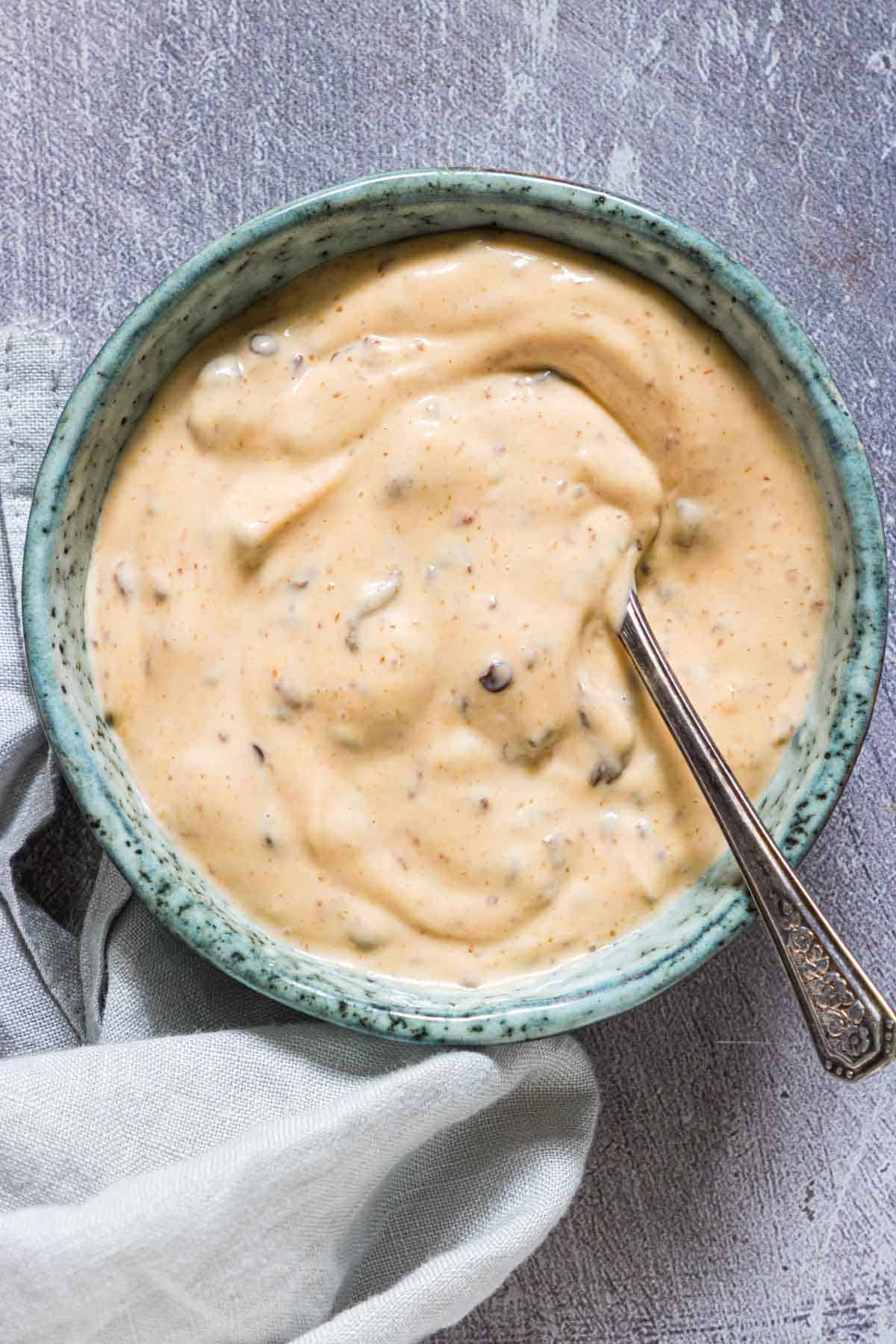 A teal bowl filled with creamy, speckled sauce, with a silver spoon resting inside. The bowl is placed on a gray textured surface next to a light gray cloth.