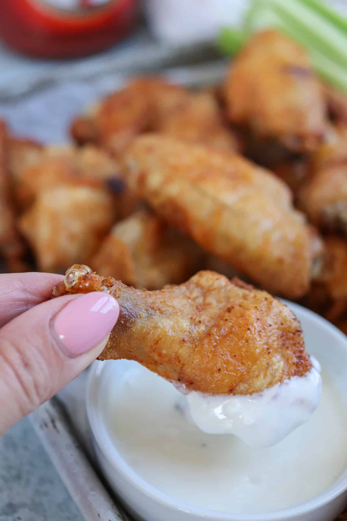 Close-up of a hand with pink nail polish holding a crispy chicken wing dipped in white sauce, one of the many delectable dipping sauces for chicken wings. In the background, a plate overflows with more wings, alongside blurred celery sticks and an inviting bowl.