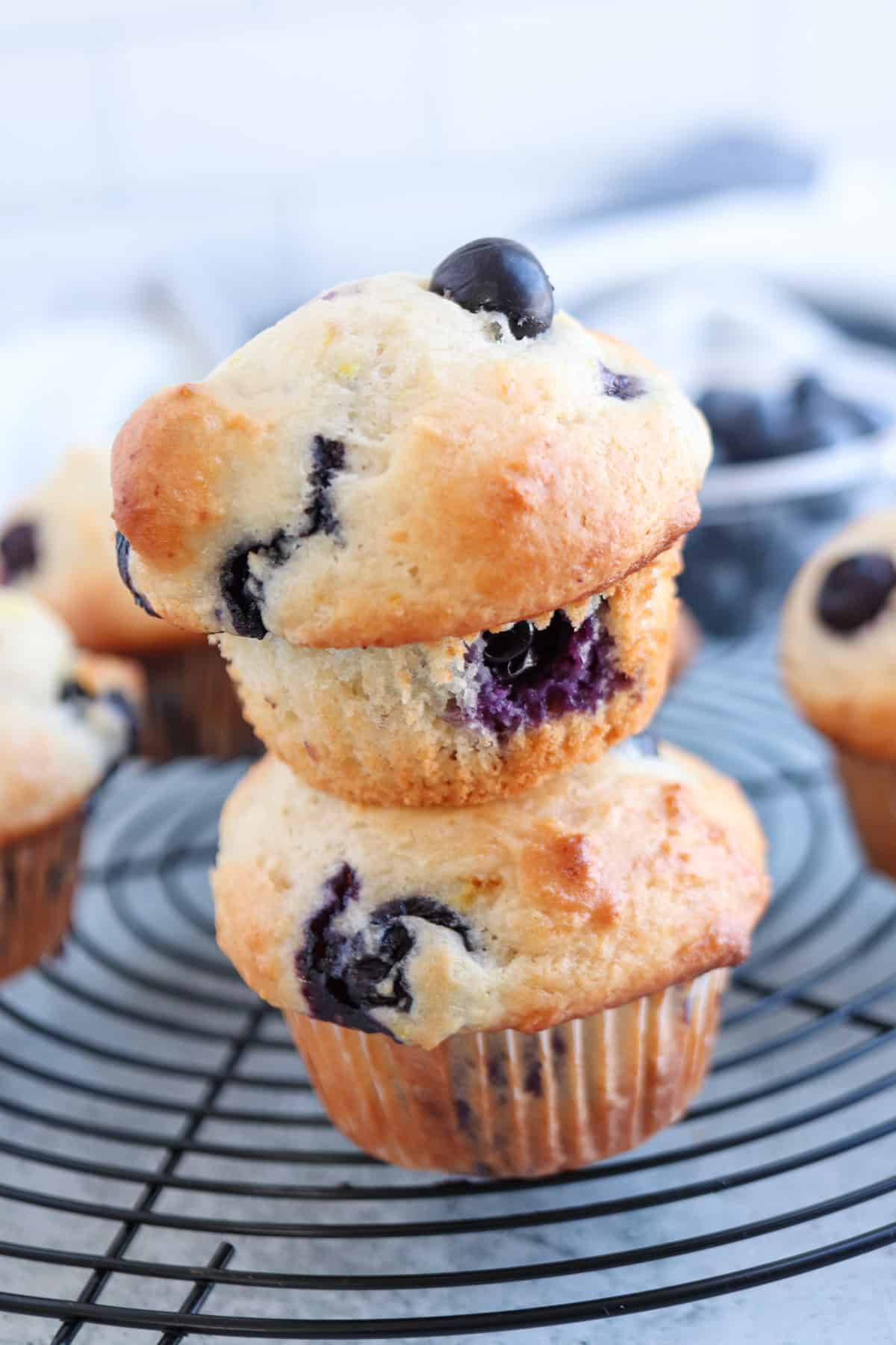 Two stacked homemade blueberry muffins with golden tops and visible blueberries, resting on a cooling rack. A bowl of fresh blueberries is blurred in the background. The scene has a cozy, rustic kitchen vibe.