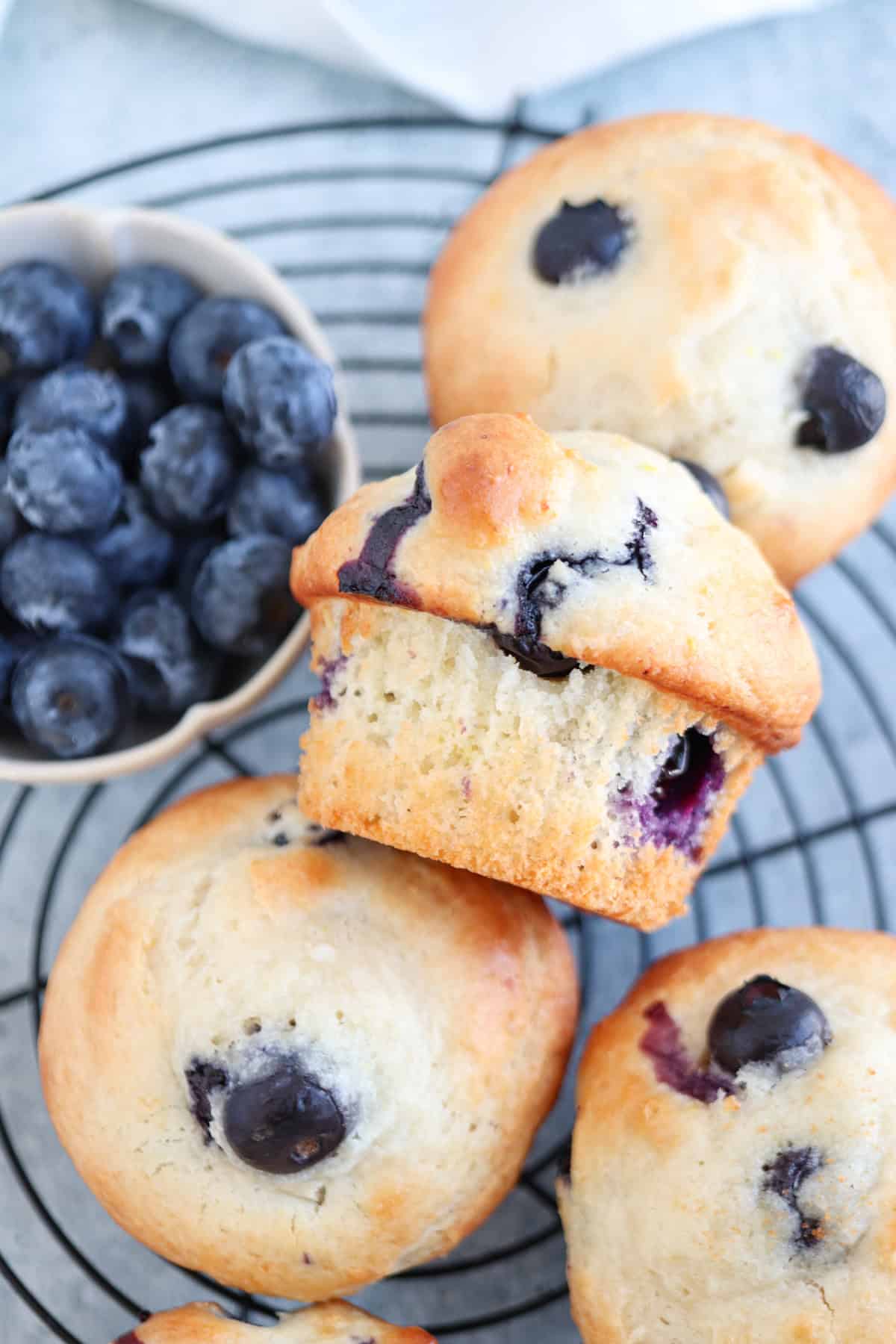 Freshly baked blueberry muffins sit on a cooling rack. One muffin is slightly tilted to show its inside. A small bowl of fresh blueberries is placed nearby. The muffins are golden with visible blueberries baked inside.