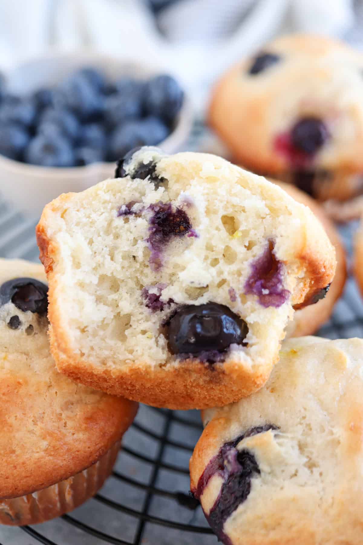 Close-up of blueberry muffins on a cooling rack. One muffin is cut in half, revealing a fluffy interior with juicy blueberries. In the background, theres a small bowl of fresh blueberries.