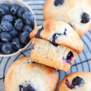 Close-up of three blueberry muffins with visible berries on top, placed on a wire cooling rack. A small bowl filled with fresh blueberries is in the upper left corner. The muffins appear slightly golden and freshly baked.