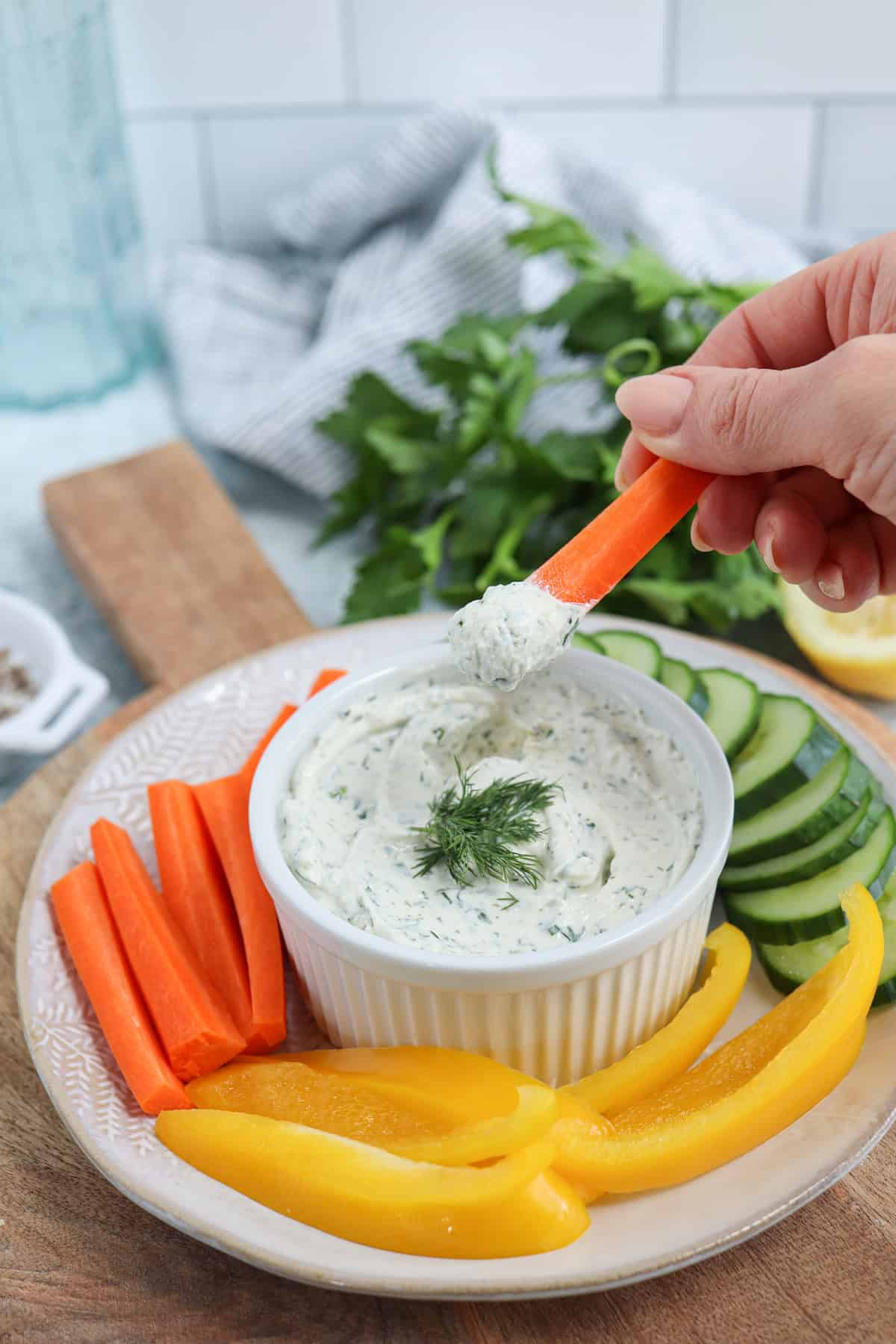 A hand dips a carrot stick into a bowl of creamy dip garnished with dill on a plate. Surrounding the dip are sliced cucumbers, carrot sticks, and yellow bell pepper slices. Fresh parsley and a striped cloth are in the background.