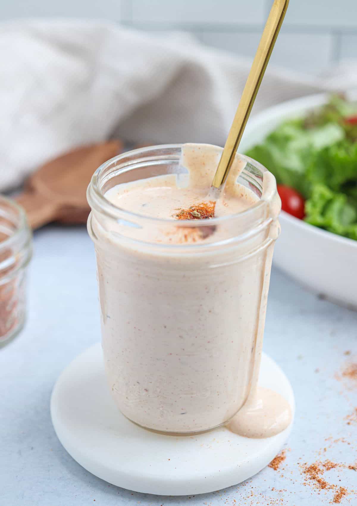 A glass jar filled with creamy dressing sits on a white coaster. The dressing is slightly overflowing, and a spoon is inside the jar. A pinch of seasoning is on top. In the background, theres a bowl of fresh salad.