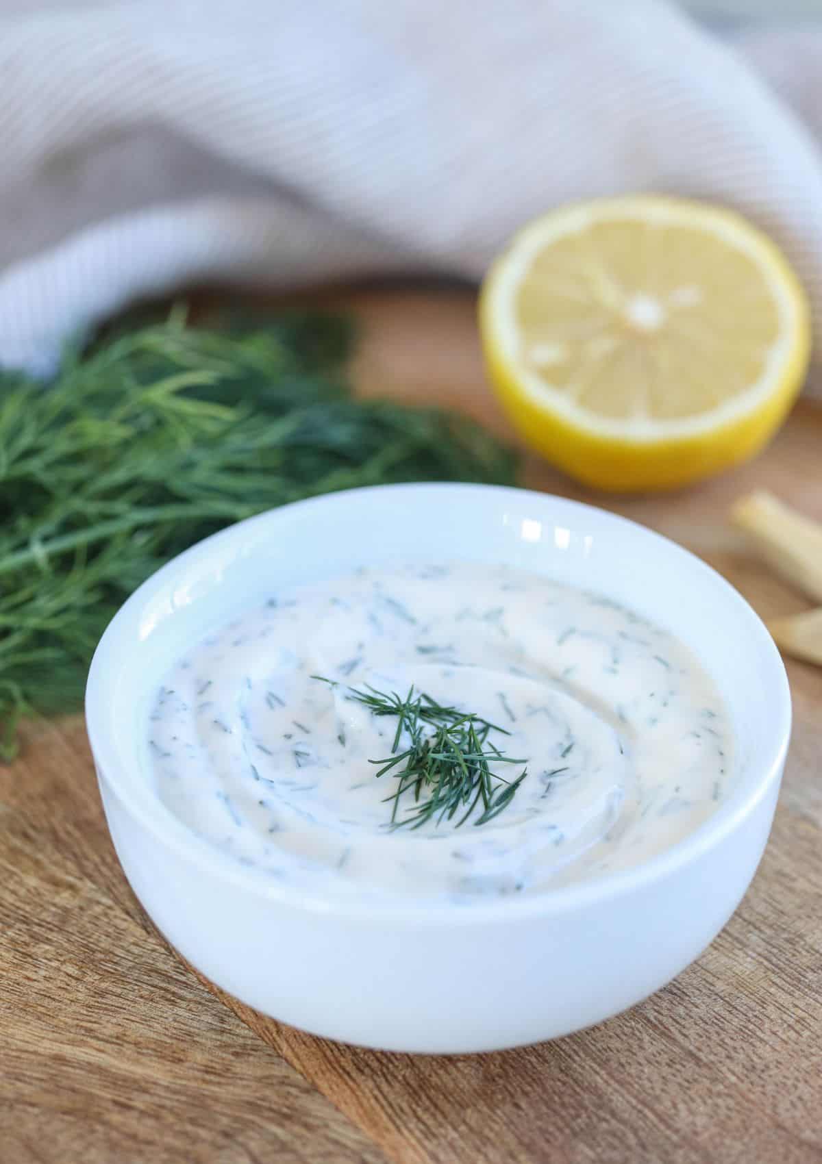 A white bowl filled with creamy dill sauce garnished with fresh dill sits on a wooden surface. In the background, there are sprigs of dill and half a lemon, along with a striped cloth.
