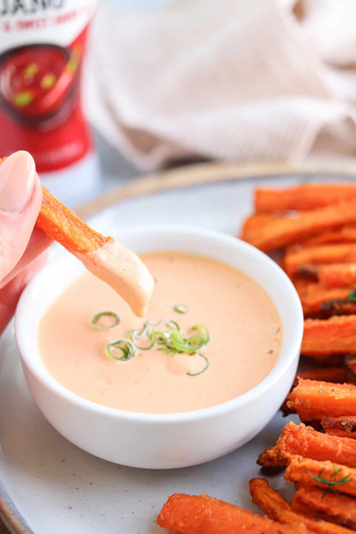 A hand dipping a sweet potato fry into a creamy sauce garnished with green onions in a white bowl. The fries are arranged on a plate beside the bowl. A can of dipping sauce is partially visible in the background.