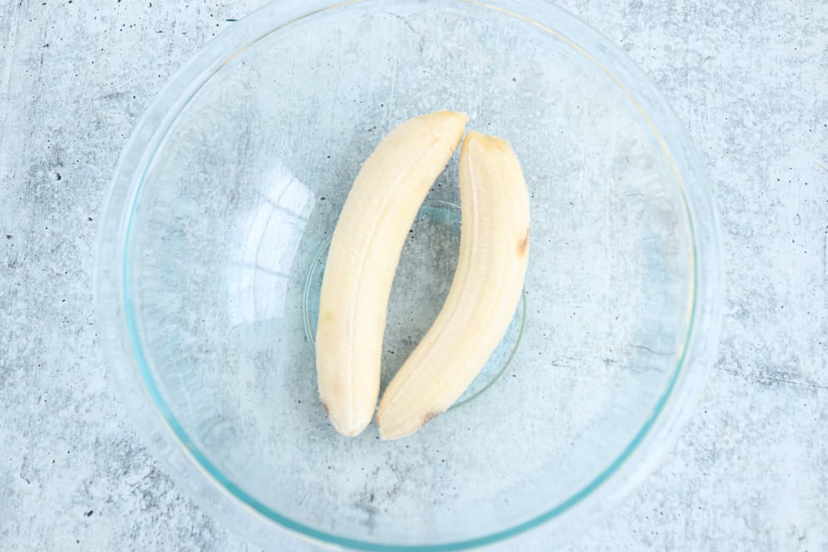 A peeled banana split in half sits in a clear glass bowl on a speckled gray surface.