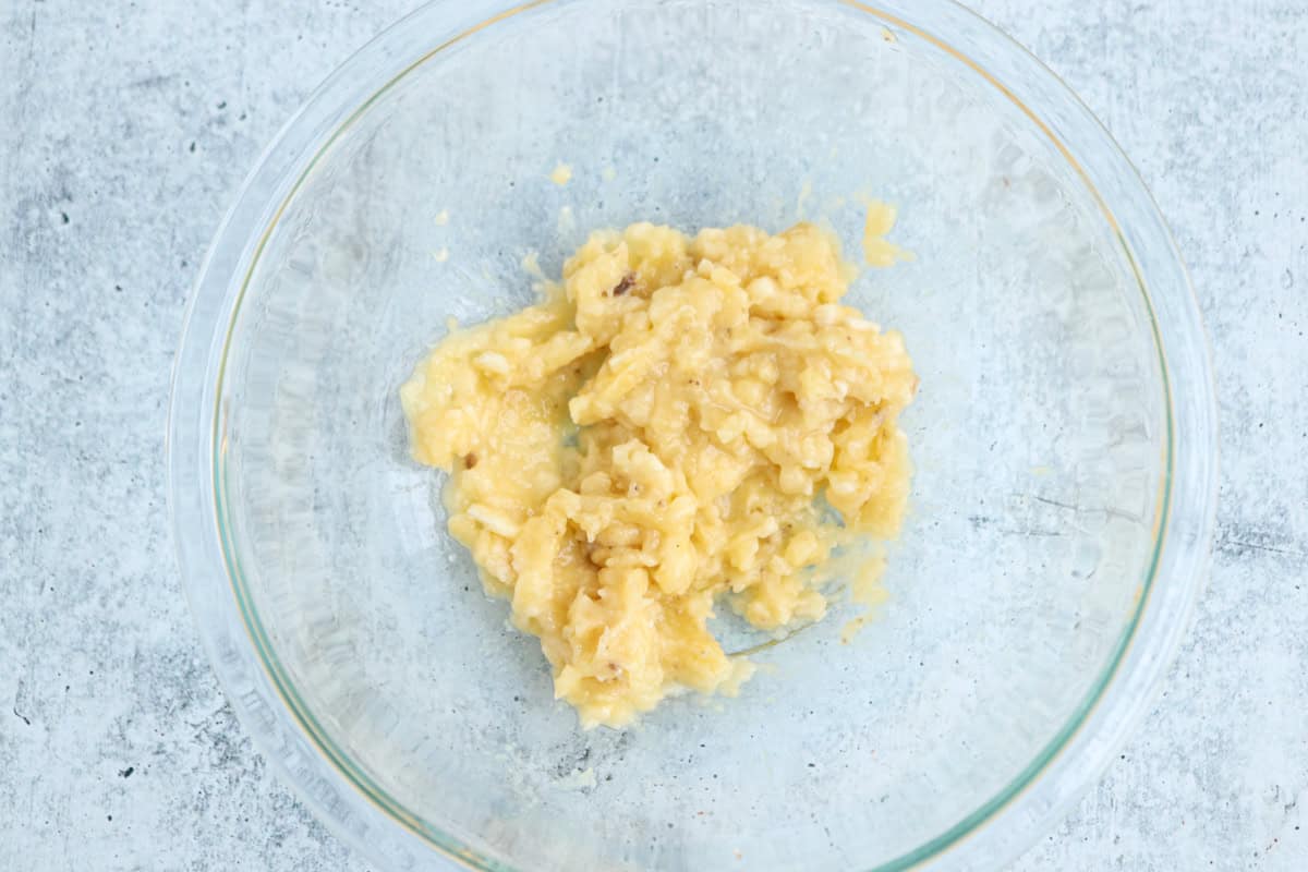 A clear glass bowl contains mashed bananas on a textured grey surface. The bananas are soft and slightly chunky, with some darker specks visible, suggesting ripeness.