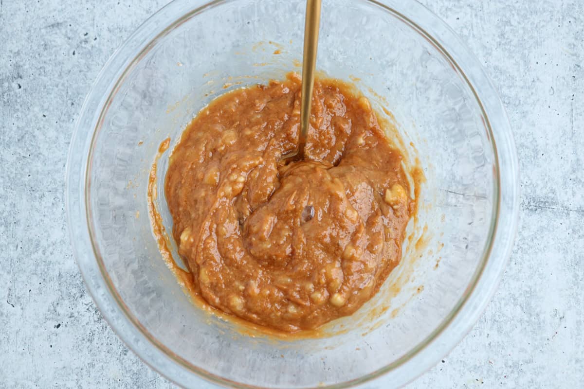A clear glass bowl contains a thick, brown batter being mixed with a fork. The batter has a slightly lumpy texture and is set on a gray, textured surface.
