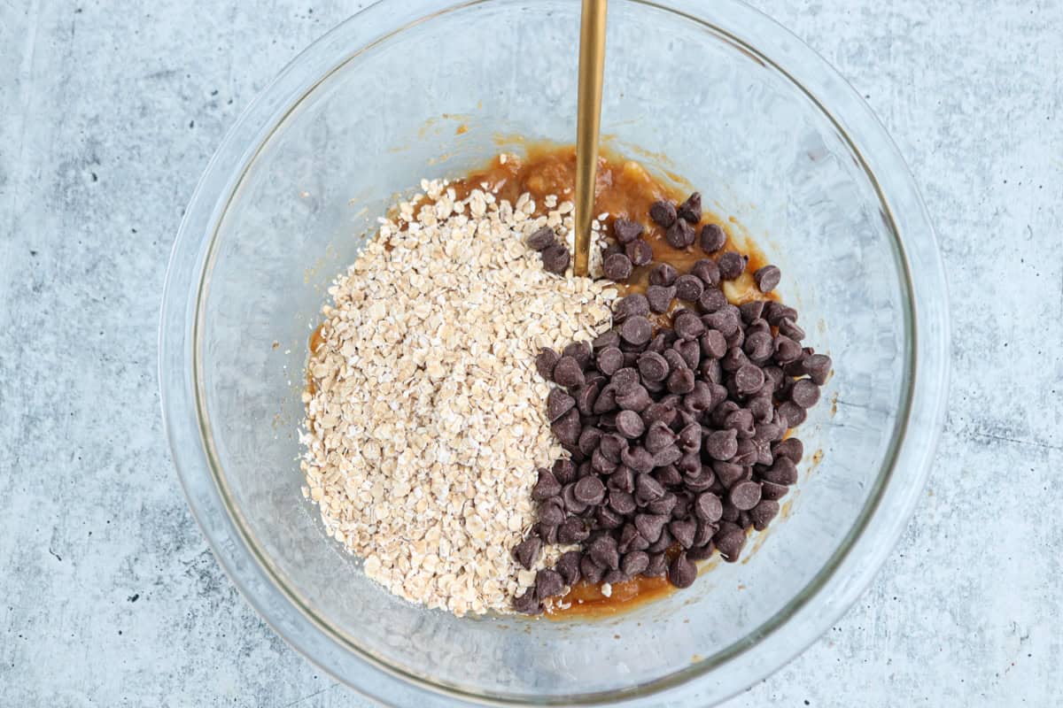 A clear glass bowl containing a mixture of oats, chocolate chips, and a brown liquid being poured. The ingredients are partially mixed on a gray textured surface.
