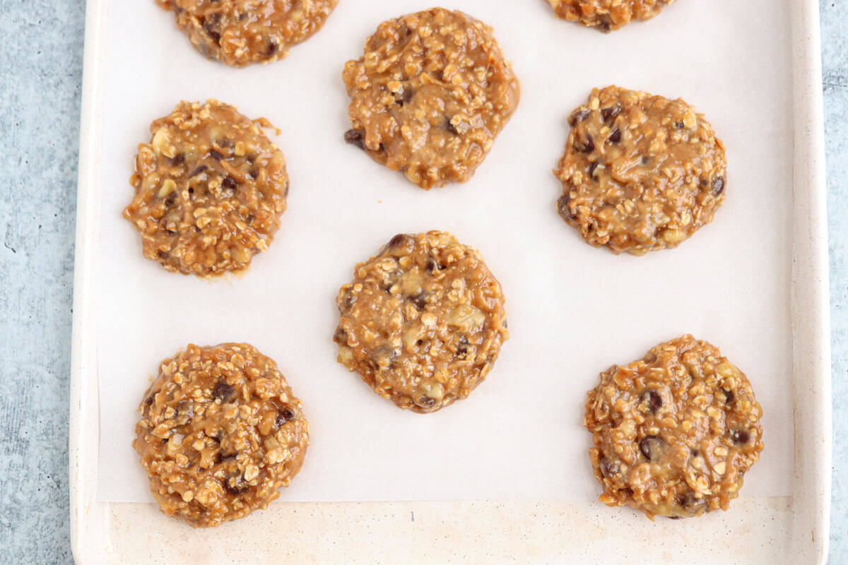 Eight no-bake cookies with oats and chocolate chips on parchment paper arranged on a baking sheet.
