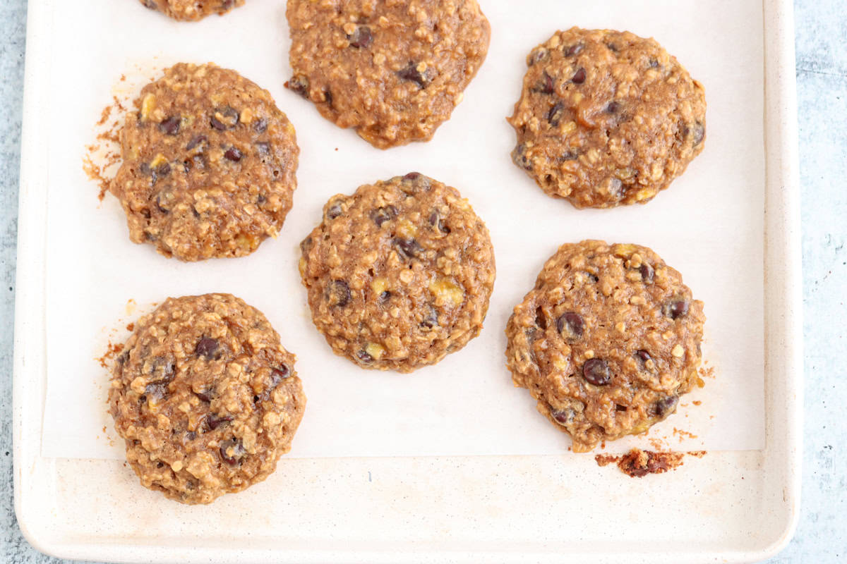 A baking sheet with seven homemade cookies featuring oatmeal and chocolate chips. They are placed on parchment paper, evenly spaced. The cookies have a rustic, slightly uneven appearance and a golden-brown color.