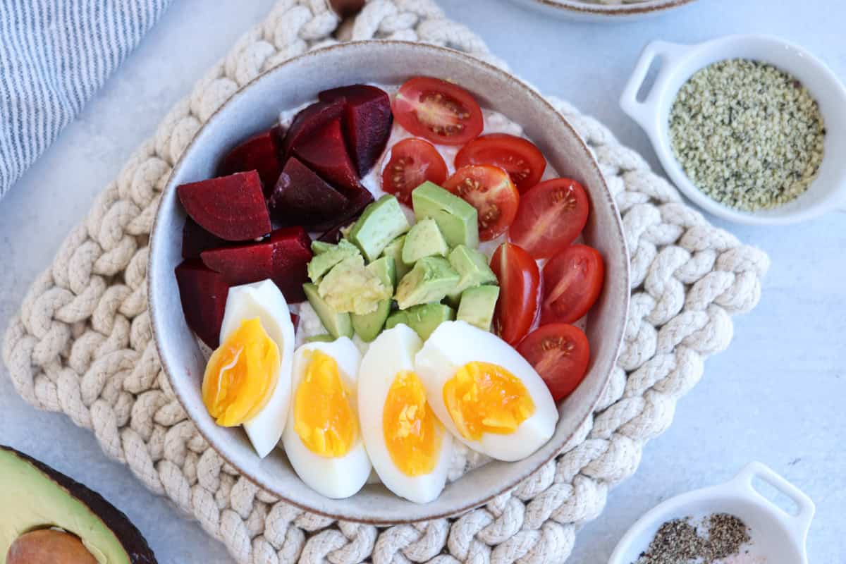 A cottage cheese breakfast bowl on a woven mat features sliced boiled eggs, diced avocado, halved cherry tomatoes, and chopped beets. Nearby are small bowls with seeds and spices, alongside half an avocado.