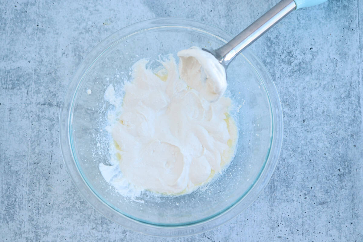 A glass bowl contains whipped white cream with a hand blender resting on the edge. The background is a textured, light gray surface.