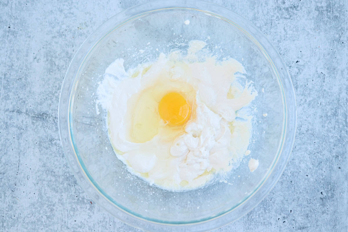 A clear glass bowl on a gray countertop contains ingredients: an egg, flour, milk, and other baking ingredients ready to be mixed. The egg is centered in the mixture.