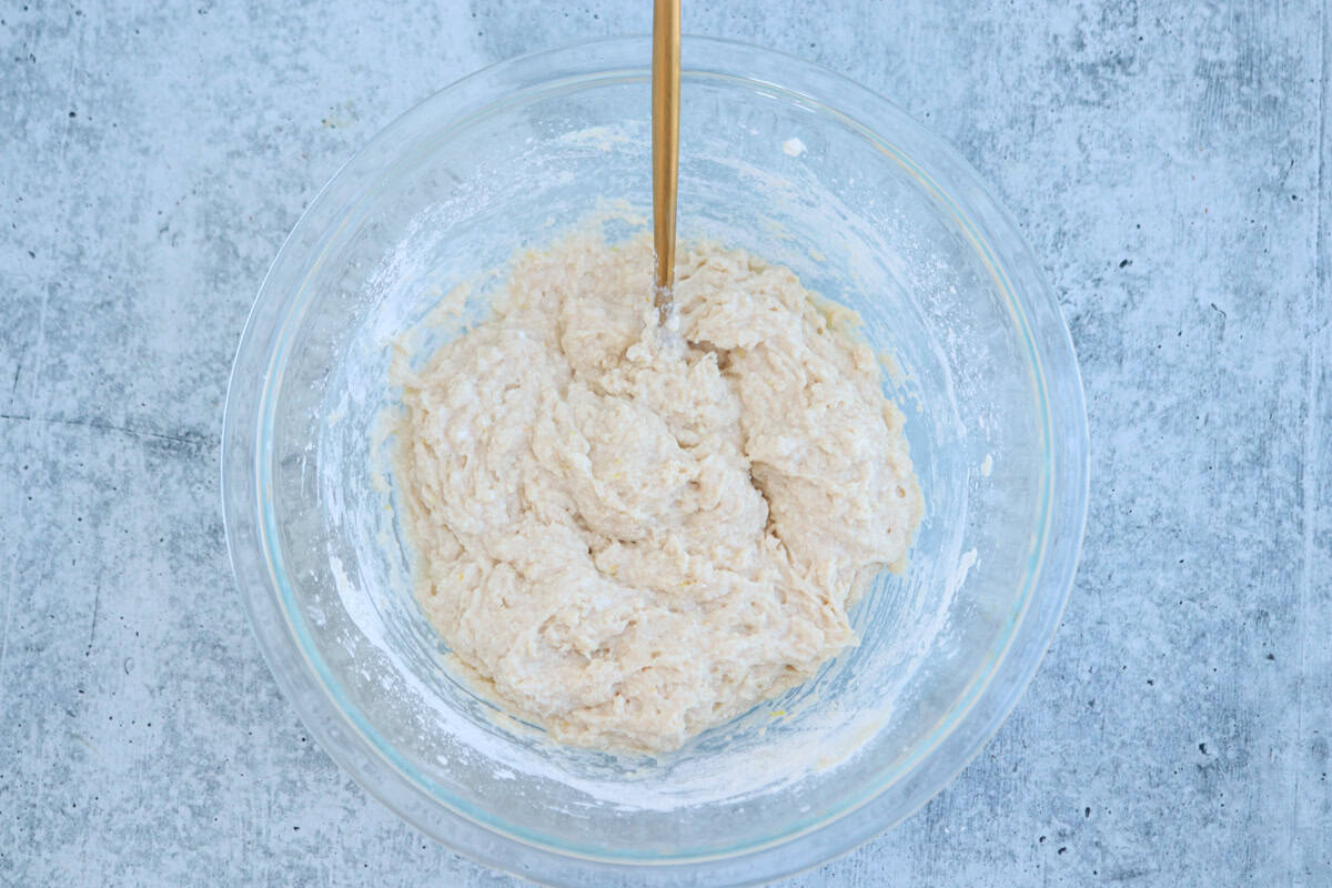A glass bowl filled with a thick, creamy dough mixture sits on a textured gray surface. A wooden spoon is partially submerged in the mixture, suggesting it is being stirred or mixed.