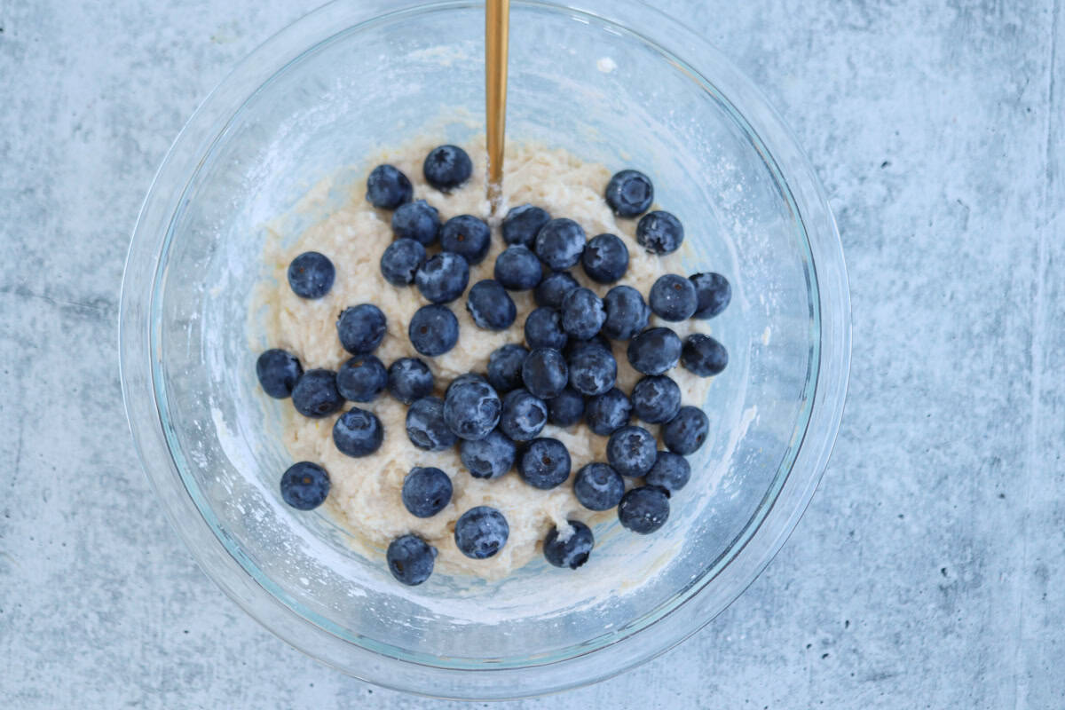 A clear glass bowl contains muffin batter mixed with fresh blueberries. A metal spoon is partially visible, embedded in the batter. The bowl is set on a gray textured surface.